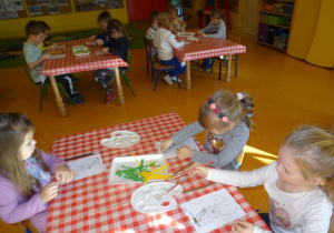 Trójka dzieci siedzi przy stoliku, wykleja wydzieranymi kawałkami papieru sylwetę Syrenki Warszawskiej.